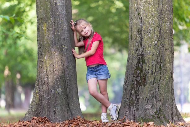 一个漂亮的小女孩的肖像站在大树干在夏季公园户外。图片下载
