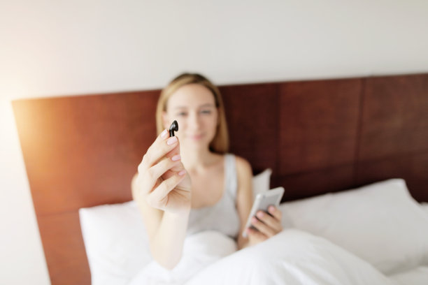 在阳光明媚的日子里，年轻女子躺在床上，专注于她手中的无线耳机图片下载