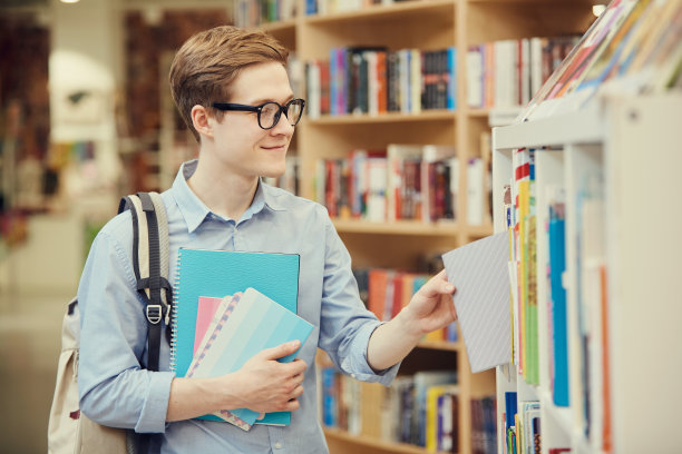 面带微笑的英俊的学生男孩戴着眼镜站在书架上，在书店选购课本图片下载