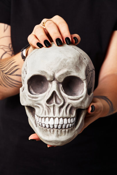 一名身穿黑色衣服、戴着黑色手表的女子的纹身手拿着一个带有牙齿的真人头骨模型。特写镜头图片下载