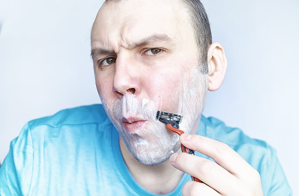 一个男人对着镜子刮胡子。剃须泡沫。一次性剃须刀。图片下载