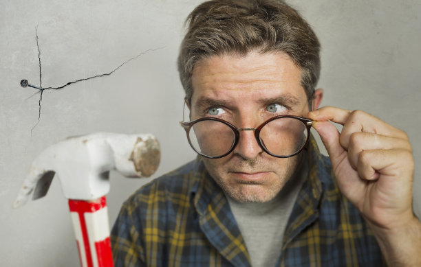 这是一个宅男在钉完钉子后想要用锤子砸挂框架的照片，他为自己DIY的灾难和家里乱七八糟的维修工作而做鬼脸图片下载