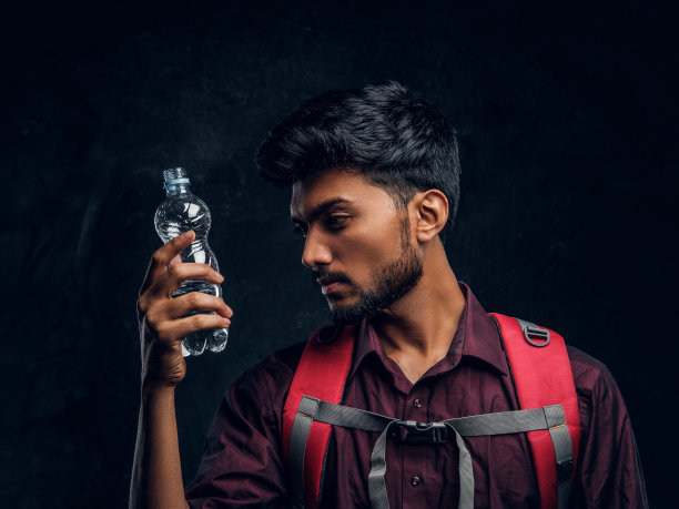 一个背包里拿着一瓶水的印度徒步旅行者看着她。工作室照片对着深色纹理的墙图片下载