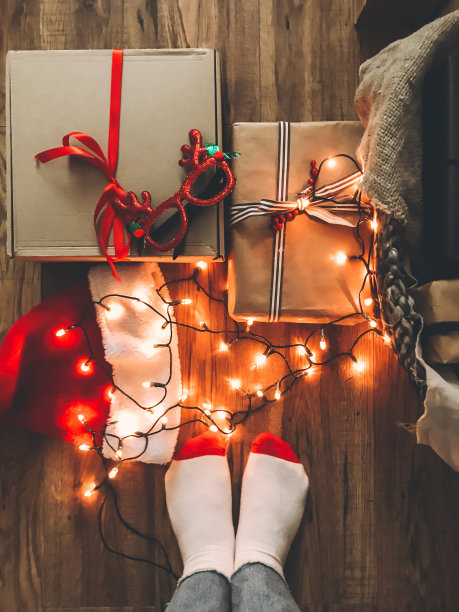 圣诞礼盒，节日灯，圣诞帽和女孩的腿在舒适的袜子在木地板在平安夜。节日快乐。前视图。手机照片图片下载