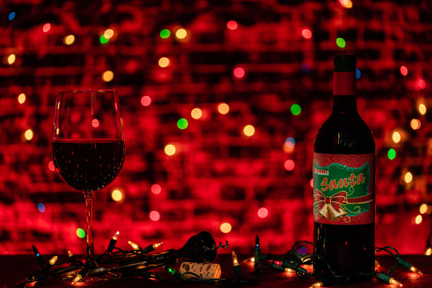一个温暖的黑暗灯光的场景，一个杯子和一瓶葡萄酒准备节日庆祝。唯一的灯光是圣诞彩灯，发出温暖的光芒。在中心背景的墙上有写字的空间。图片下载