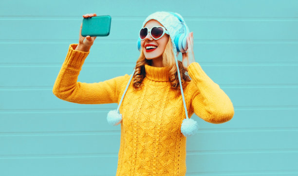 冬天的照片微笑的女人正在用手机自拍，戴着无线耳机，穿着黄色针织毛衣，白色帽子，蓝色背景的心形太阳镜听着音乐图片下载