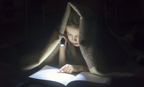 女孩晚上拿着手电筒在毯子下看书。图片下载