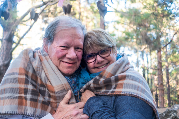 一对相爱的老年情侣在毯子下互相拥抱取暖。微笑着看着镜头。背景是一片松树林图片下载