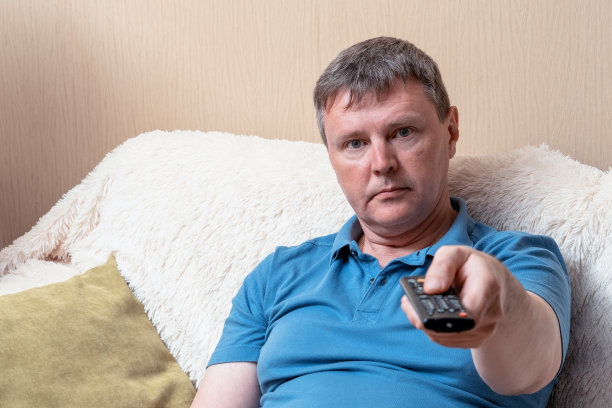 穿着蓝色t恤的中年白人拿着电视遥控器。一个坐在舒适沙发上的男人打开了电视频道图片下载