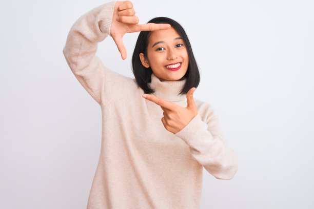 年轻的中国妇女穿着高领毛衣站在孤立的白色背景微笑做框架与手和手指与快乐的脸。创意和摄影概念。图片下载