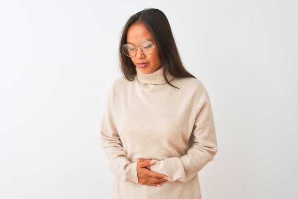 年轻的中国妇女穿着高领毛衣和眼镜在孤立的白色背景下，手在胃上，因为恶心，痛苦的疾病感觉不舒服。疼痛的概念。图片下载
