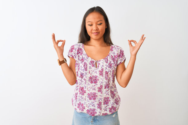 年轻的中国女人穿着休闲花t恤站在孤立的白色背景放松和微笑，闭上眼睛做冥想手势与手指。瑜伽的概念。图片下载