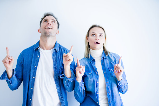 年轻美丽的夫妇穿着牛仔衬衫站在孤立的白色背景惊讶和惊讶的看着，用手指和举起的手臂。图片下载