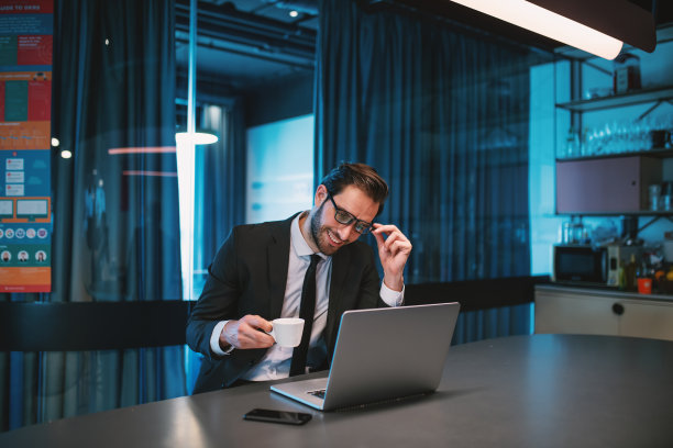 穿着西装、戴着眼镜、满脸胡须、面带微笑的白人商人，端着一杯刚煮好的咖啡，坐在公司厨房里看笔记本电脑。图片下载