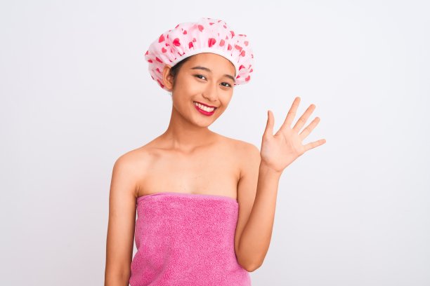 年轻的中国女子穿着浴巾和浴帽，在孤立的白色背景下，用第五根手指指着前方，自信而快乐地微笑着。图片下载