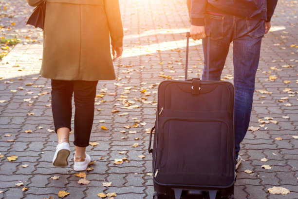 一个女人和一个男人拿着行李箱在阳光明媚的早晨走在街上的秋天，而去机场旅游度假旅行在一个现代城市背景的近背影视图图片下载