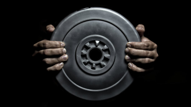 在健身房里，一名身穿蓝绿色t恤和黑色裤子的年轻女子从黑色背景的吧台上举起煎饼图片下载