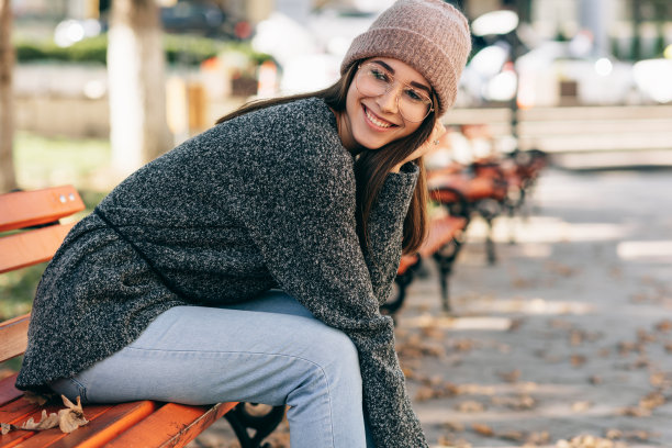 这张照片里的女学生面带微笑，看着镜头，穿着针织毛衣，戴着眼镜和帽子，坐在城市街道的长凳上。漂亮的年轻女子在外面休息图片下载