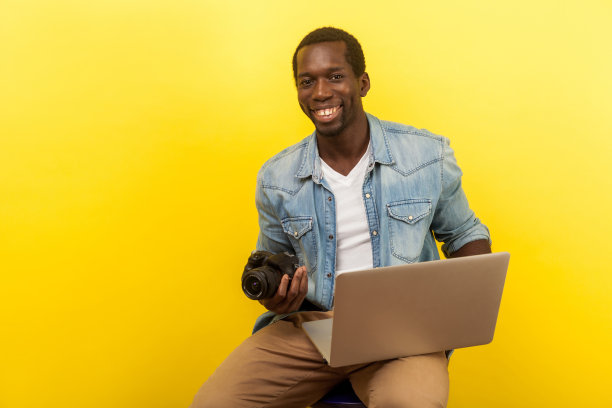 肖像的精力充沛的正摄影师持有数码单反相机和笔记本电脑。孤立在黄色背景上图片下载