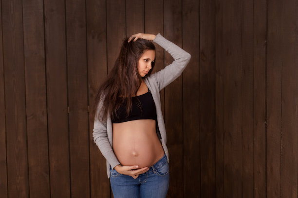 穿着牛仔裤和黑色上衣的孕妇双手搭在深棕色的腹部上。怀孕、分娩、准备和期望概念。美丽柔情的怀孕照片图片下载
