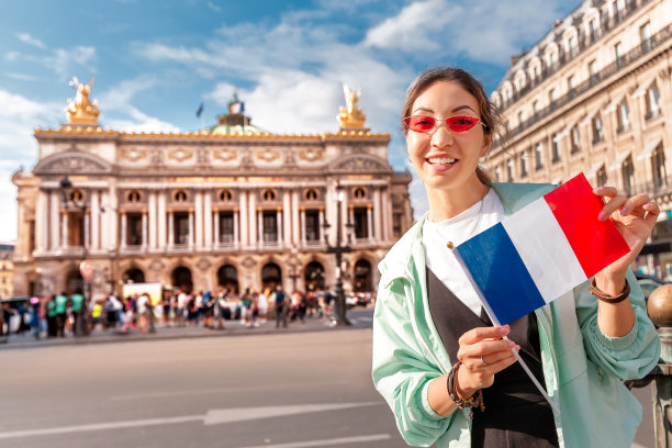 在巴黎音乐学院历史建筑的加尼埃歌剧院的主立面附近，快乐的亚洲女孩旅行者举着法国国旗。法国景点和文化景点的概念图片下载