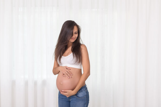 穿着牛仔裤和白色上衣的孕妇双手放在白色背景下的肚子上。怀孕、分娩、准备和期望概念。美丽柔情的怀孕照片。图片下载