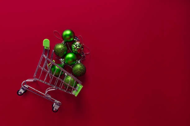 红色背景上放着一辆从超市买来的小金属手推车，里面装满了绿色的圣诞玩具。玩具从篮子里得到足够的睡眠。水平的照片。交易的概念。平的。本空间图片下载