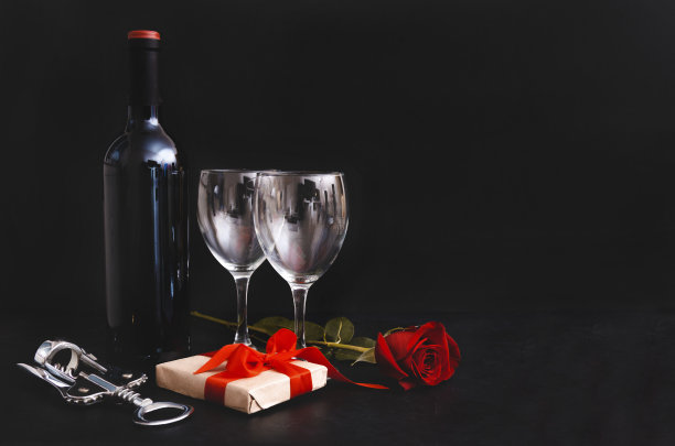 葡萄酒瓶、两个酒杯、深色葡萄、开瓶器、红玫瑰、带丝带的礼品盒在黑色背景上，留白素材图片