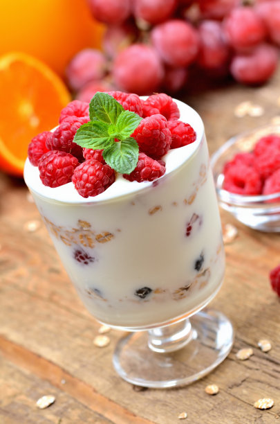 特写酸奶与燕麦和新鲜的覆盆子在一个玻璃杯，薄荷叶，橙子和葡萄在背景-垂直照片图片下载