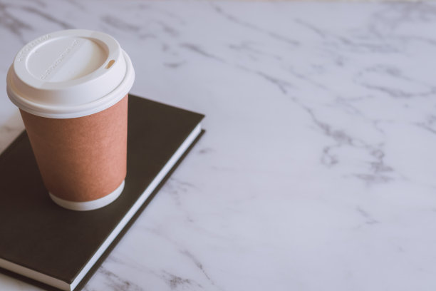棕色的纸杯咖啡，黑色的书，课本或日记，白色的大理石桌子上有复印空间。品牌和标志的照片概念，咖啡馆，休闲，阅读，放松。餐饮形象。图片下载