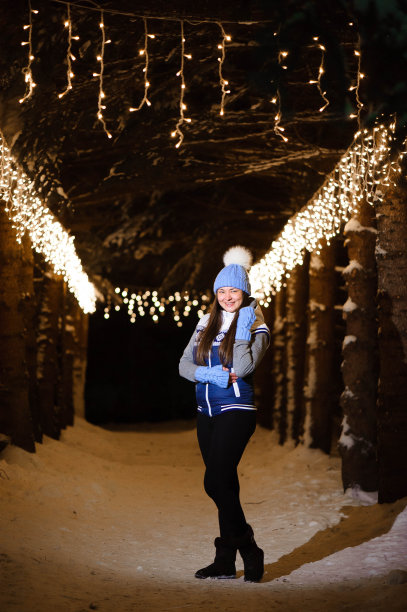 户外照片年轻美丽的快乐微笑的女孩，摆姿势在装饰着灯泡的小巷。圣诞集市的背景。图片下载