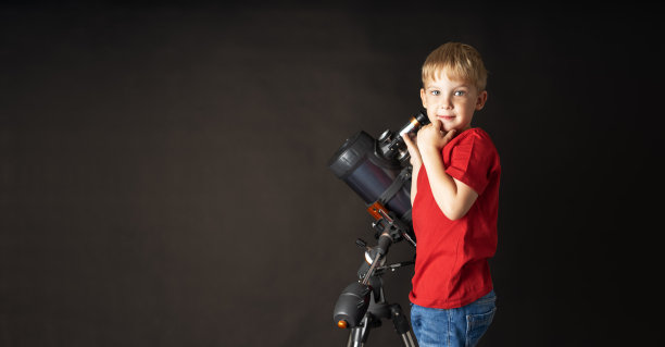 一个穿着红色足球的小男孩站在一个大望远镜旁边。图片下载