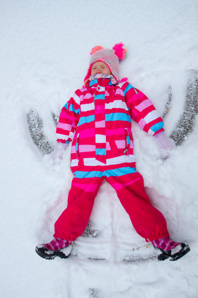 可爱的蹒跚学步的女孩穿着鲜艳的颜色，在寒冷的冬天户外做雪天使图片下载