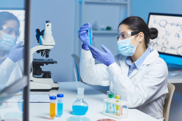 严肃的年轻印度女化学家戴着护目镜和面罩坐在实验室的办公桌前，在试管中检查蓝色液体图片下载