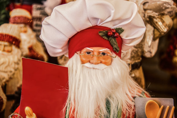 装饰圣诞娃娃在厨师帽拿着空白标志和烹饪工具-特写和选择性聚焦-其他模糊的圣诞老人在背景图片下载