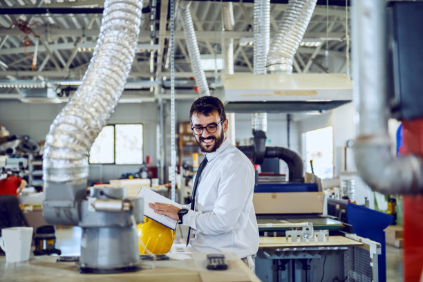 年轻有经验的白种人胡须图形工程师，戴着眼镜，拿着笔记本，在打印车间检查印刷机。图片下载