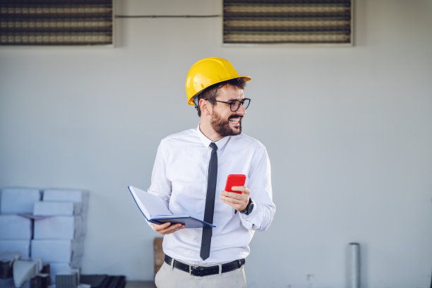 头戴头盔微笑的白人技术人员站在打印店前，拿着智能手机和笔记本电脑，看向别处。图片下载
