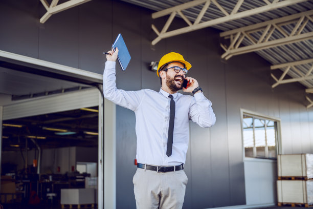 满脸胡须、头戴头盔的白种人首席执行官兴高采烈地站在印刷厂前，用智能手机交谈。客户是满意的。图片下载