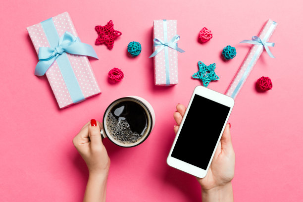 俯视图的一个女人拿着手机在一只手和一杯咖啡在另一只手在粉红色的背景。圣诞装饰品和玩具。新年假期概念。模型图片下载