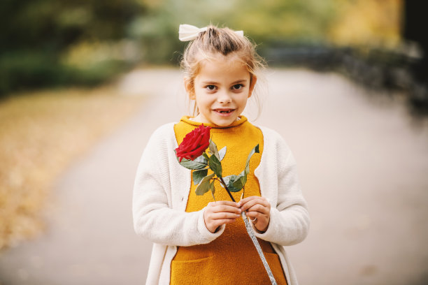 梳着马尾辫的可爱金发小女孩站在公园里，手里拿着红玫瑰。秋天的时间。图片下载
