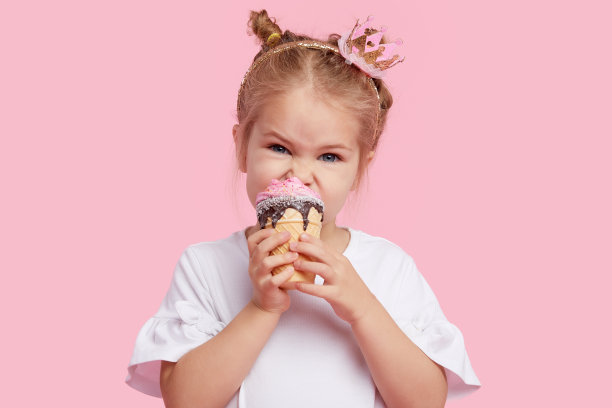 可爱的小女孩愉快地吃着美味的冰淇淋在一个粉红色的工作室背景。闭着眼睛舔。婴儿食品和快乐童年的概念图片下载