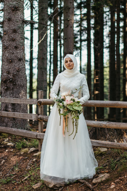 美丽优雅的新娘与希贾布在蕾丝婚纱与长长裙和长袖。她手里拿着一大束花。在户外，在树林里。图片下载