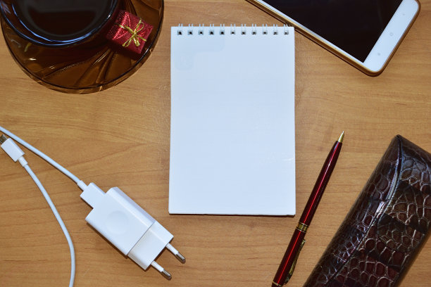 桌上空笔记本，周围是钢笔、电话、玻璃杯、烧瓶、电话、充电器和礼物。图片下载