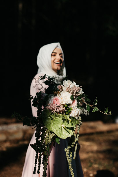 美丽优雅的新娘戴着头巾，裸粉色长裙和长袖。在户外，在路上。她在为时装摆姿势。她手里拿着一大束花。图片下载