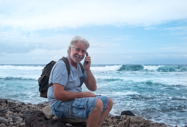 一位留着白胡子的老人坐在海滩上打电话的肖像。背景是水面上的地平线。多云的天空和海浪为冲浪爱好者图片下载