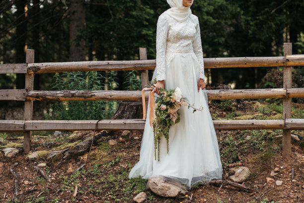 美丽优雅的新娘与希贾布在蕾丝婚纱与长长裙和长袖。她手里拿着一大束花。在户外，在树林里。图片下载