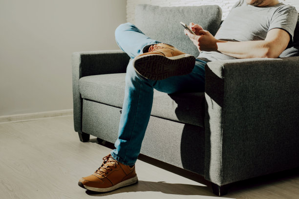 这个男人坐在一个灰色的沙发上，正在用手机。使用电话和在电话上打字的概念。发送短信，查看短信。图片下载