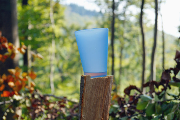 蓝色塑料杯在森林的树干或树桩上，在一个树木繁茂的自然景观背景。美丽的风景背景上的蓝色塑料杯。图片下载