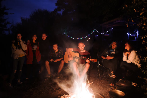 朋友旅行者在篝火边放松，听着原声音乐，在夜晚的森林里露营。一群人在篝火旁休息，在湖边露营图片下载