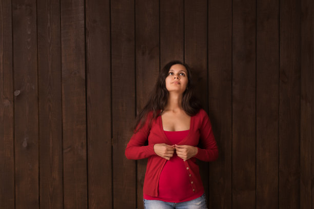 圣诞节孕妇在牛仔裤短裤和红色上衣牵手腹部在一个深棕色的背景。怀孕，母性，期望概念。美丽柔情的怀孕照片图片下载